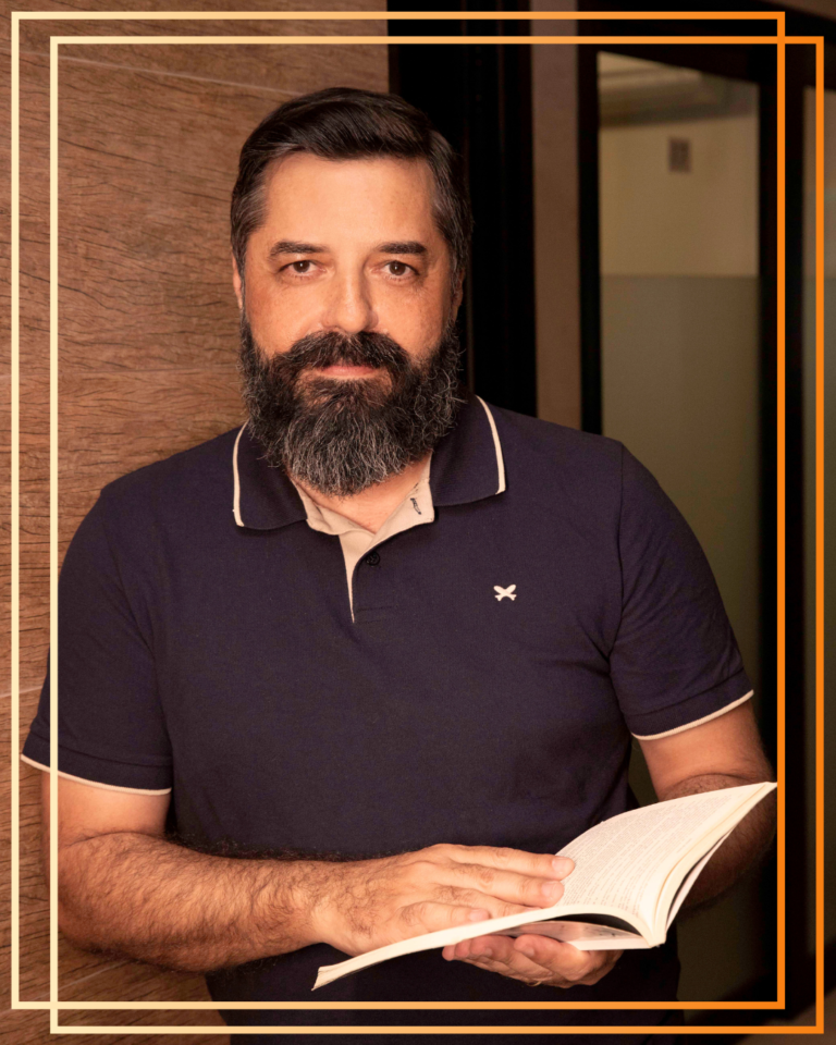 Homem com barba e cabelo escuro, vestido com uma camisola polo azul, a segurar um livro aberto e a olhar diretamente para a câmara.
