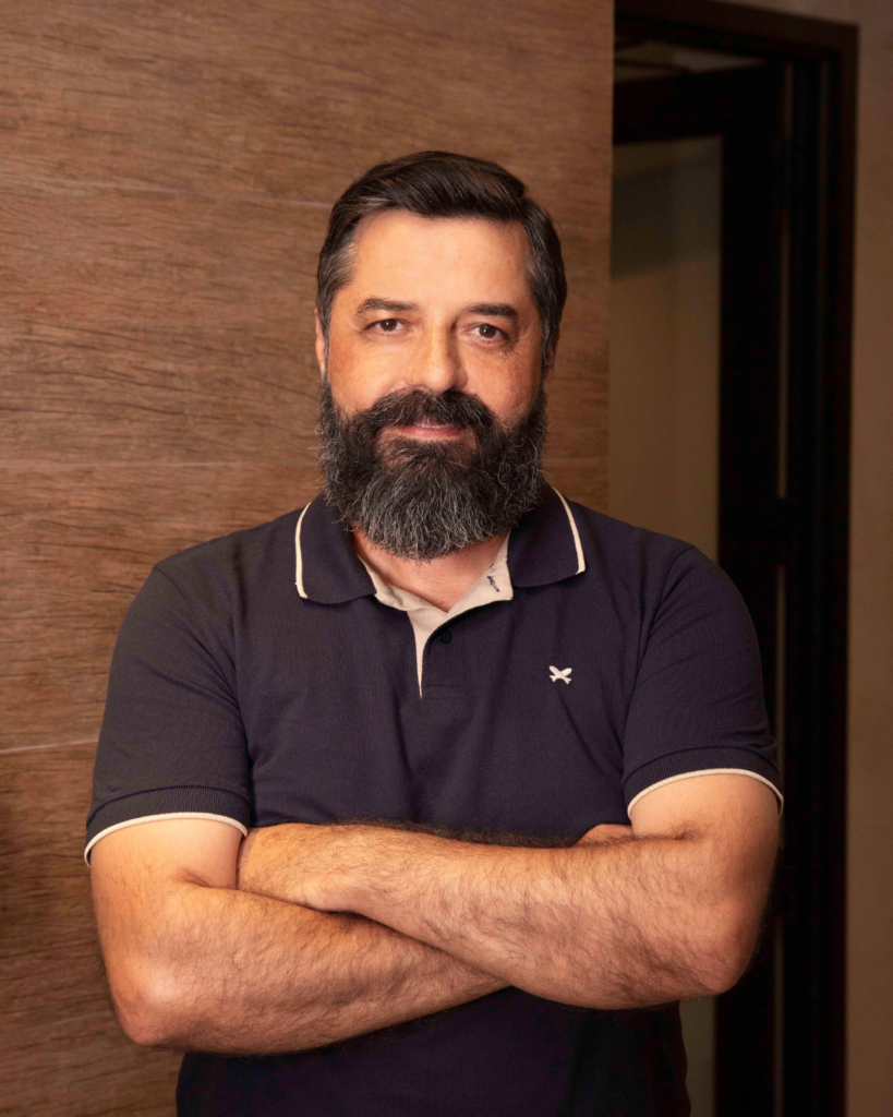 Homem com barba e cabelo escuro, vestido com uma camisola polo azul, de braços cruzados e a olhar diretamente para a câmara.