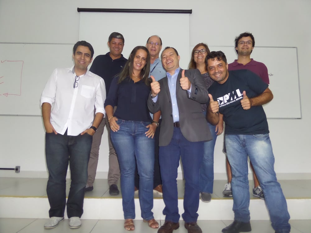 Grupo de sete pessoas a posar num palco em frente a um quadro branco e ecrã de projeção, com gestos de aprovação.