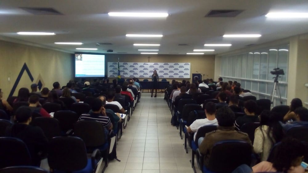 Auditório com público atento a uma apresentação, com orador em palco e ecrã de projeção ao lado.