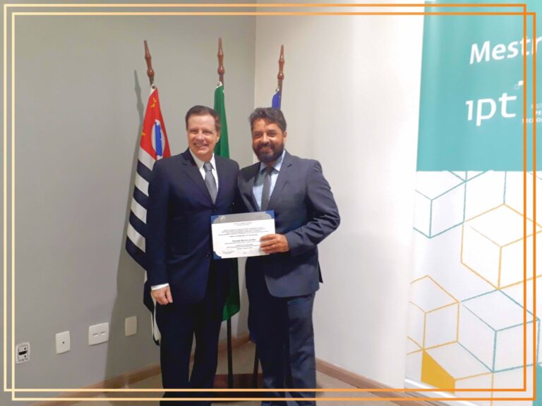 Dois homens em traje formal posam para fotografia, um deles segurando um diploma, com bandeiras do Brasil e de São Paulo ao fundo